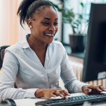 Young beautiful joyful african american woman smiling while working on a laptop in a open space office. Attractive young woman working in an coworking office with her colleagues.